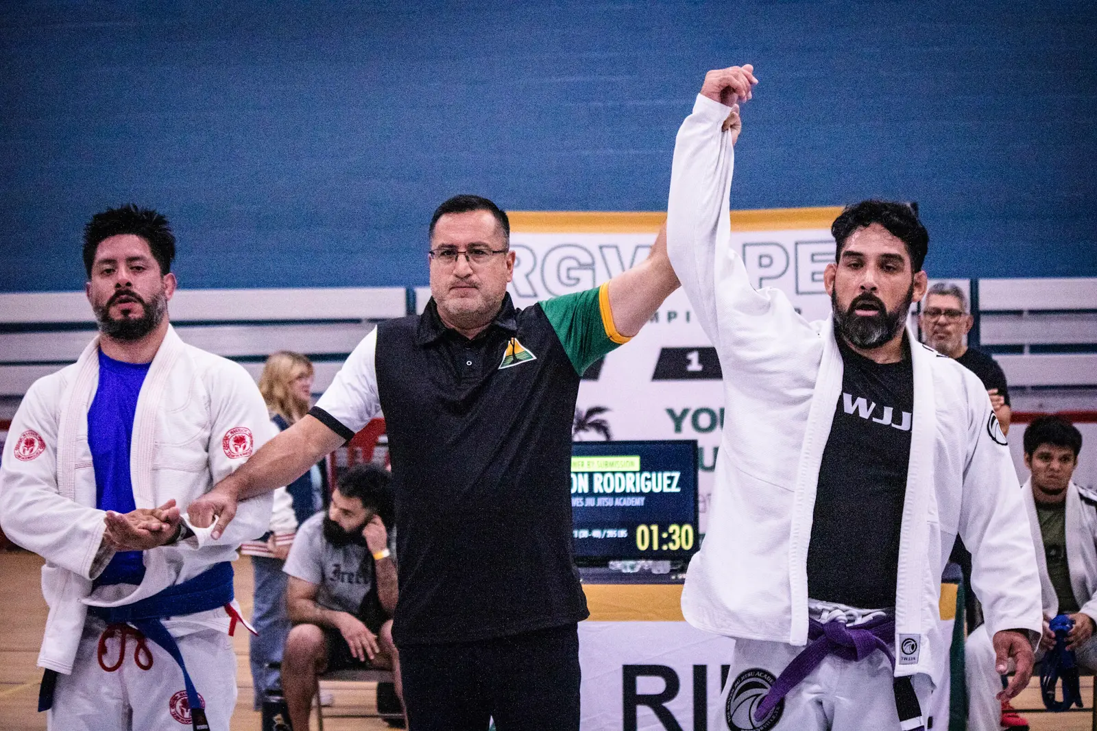 An RGV Open referee raises the winner's hand after an adult gi match, in front of the RGV Open banner and scoreboard.